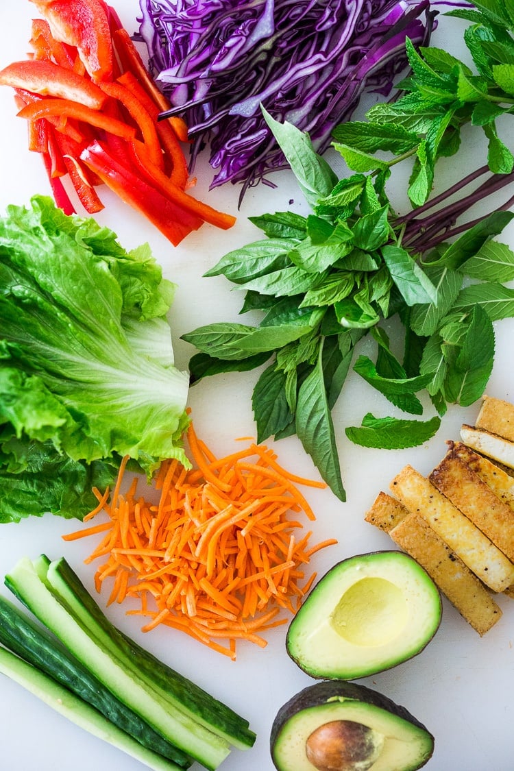 ingredients laid out for fresh spring rolls - sliced cabbage, red bell pepper, fresh herbs, lettuce, baked tofu, shredded carrots, cucumber, and avocado.