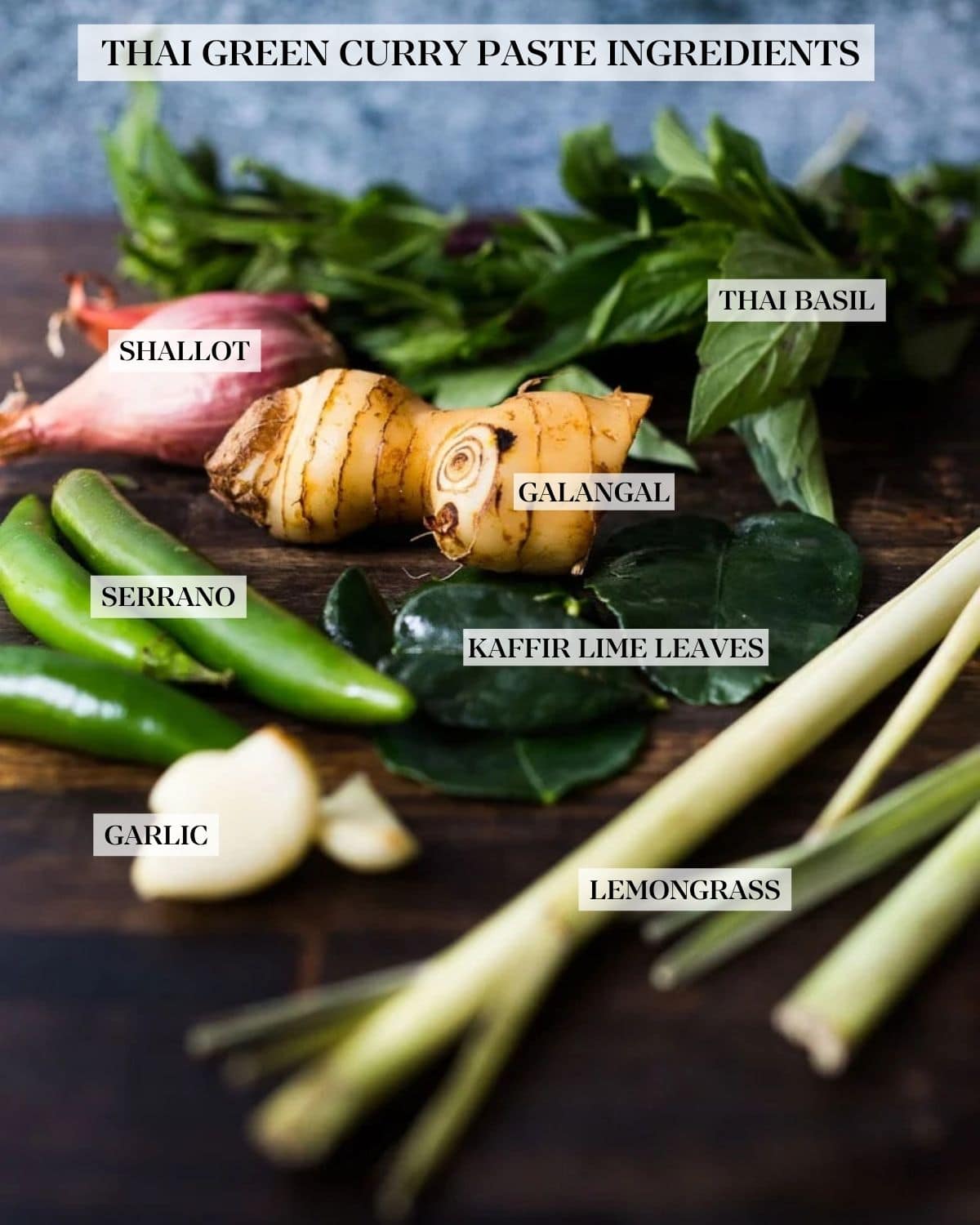 Thai green curry paste ingredients laid out on a wood table and labeled - shallot, thai basil, galangal, serrano, kaffir lime leaves, garlic, and lemongrass.
