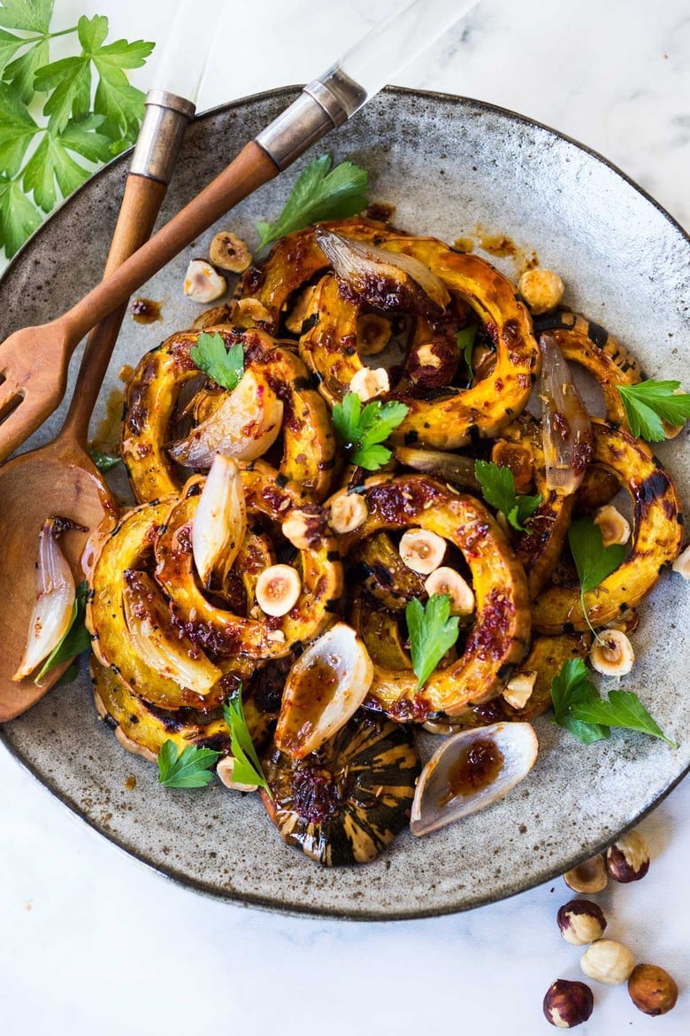 This Roasted Delicata Squash recipe is so flavorful! Made with shallots, toasted cumin seeds, and a Maple Apple Cider Glaze, topped with toasted hazelnuts. Tangy, earthy, sweet and spicy, a tasty fall side dish.