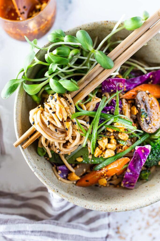 bowl of sesame noodles with mushrooms, purple cabbage, peppers, broccoli, and sprouts, with chopsticks on top and noodles twirled around. 