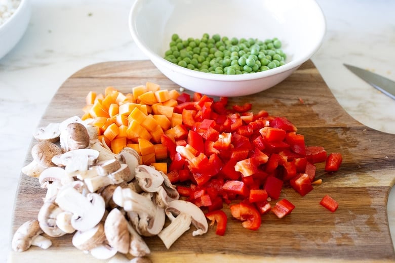 chopped mushrooms, peppers, carrots on wood cutting board next to bowl of peas.