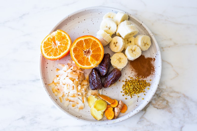 Toasted Coconut Smoothie Ingredients on a plate including: banana, dates, coconut, turmeric , ginger cinnamon, orange and bee pollen. 