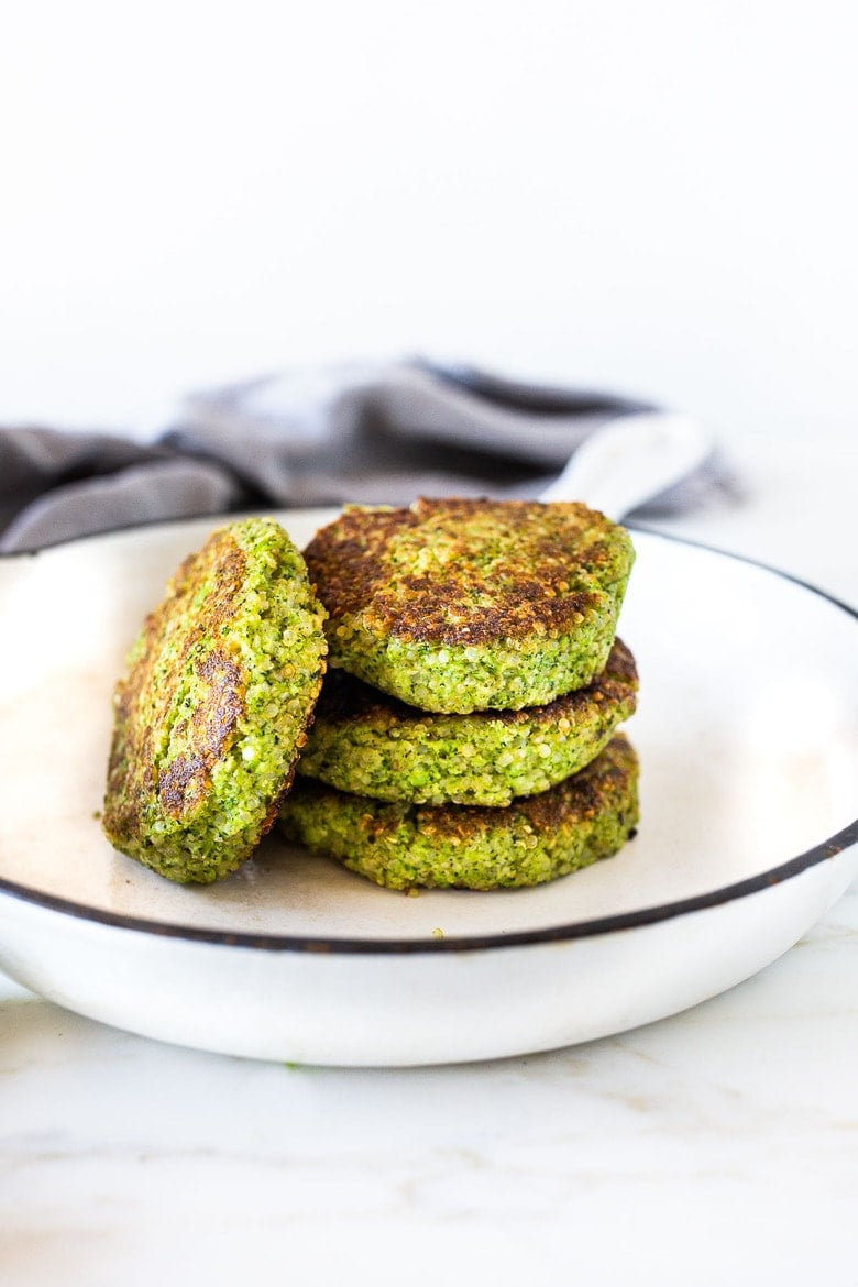 stack of broccoli quinoa cakes with one sideways, showing seared sides and crisp quinoa edges, on white serving dish.