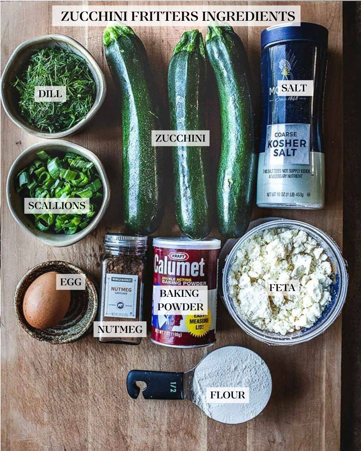 ingredients for zucchini fritters laid out on wood board - kosher salt, zucchini, dill, scallions, egg, nutmeg, baking powder, feta, and flour.
