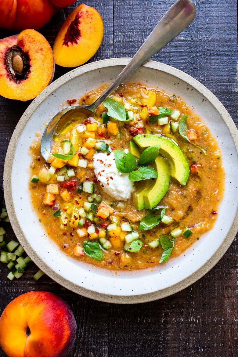 Tomato Peach Gazpacho with basil and burrata and sliced avocado in bowl with spoon.