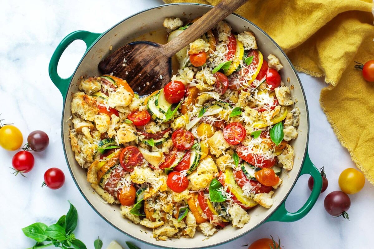 round baking dish with tomato zucchini gratin with wood serving spoon, blistered cherry tomatoes, and grated cheese. 