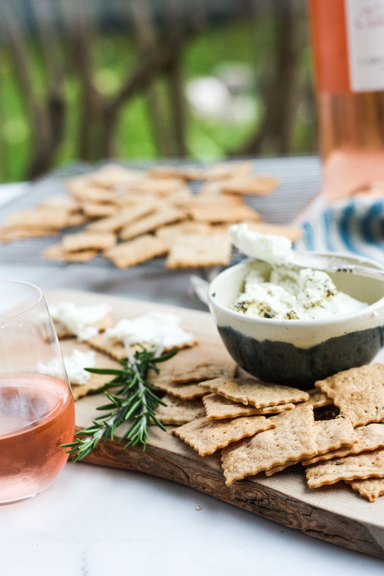 pile of homemade sourdough crackers on wood serving board alongside dip and glass of rose wine with fresh rosemary for garnish.