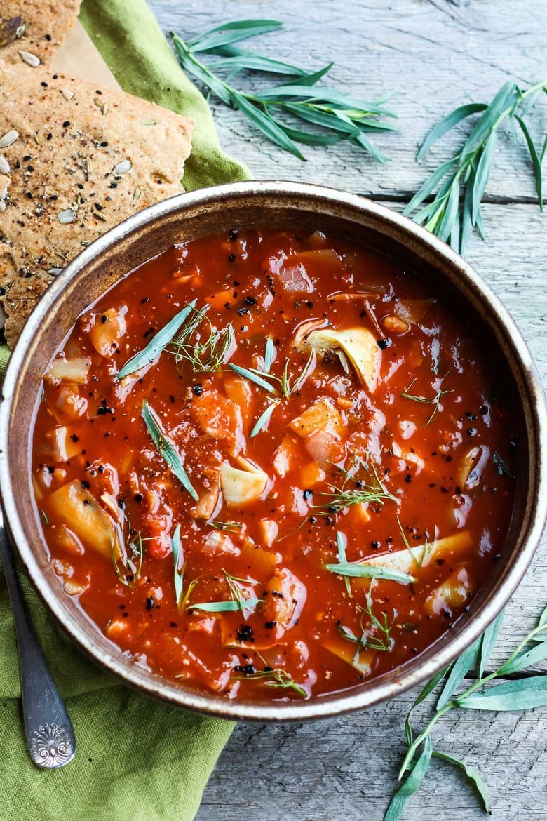 tomato artichoke soup with fennel and tarragon in bowl with colorful veggies and fresh herbs.