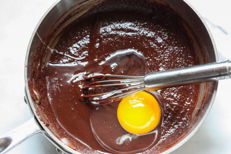 Chocolate mixture in sauce pan with a whisk and an egg cracked into it.