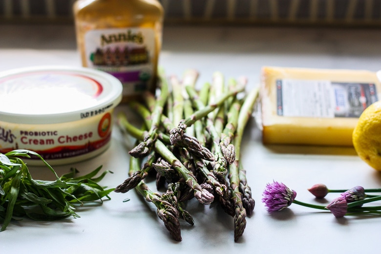 ingredients for asparagus tart laid out on counter- gruyere cheese, chive blossoms, asparagus bunch, cream cheese, French tarragon, dijon mustard, lemon.