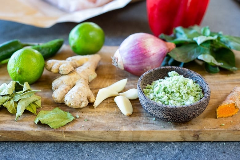 thai fish curry ingredients - wood board with turmeric, green curry paste, garlic, lemongrass, ginger, kaffir lime leaves, limes, jalapeno peppers, shallot, and basil.