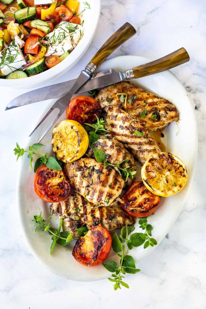 serving platter with grilled chicken, grilled tomatoes, and grilled lemons, topped with herbs.