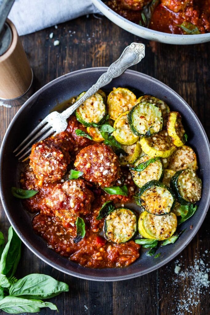 bowl of sun-dried tomato meatballs with baked zucchini, garnished with parmesan and basil, with fork. 