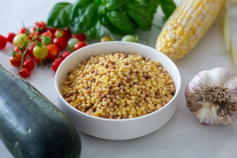 Ingredients for Fregola Sarda on counter - fregola in bowl, garlic head, corn, basil, cherry tomatoes, zucchini.