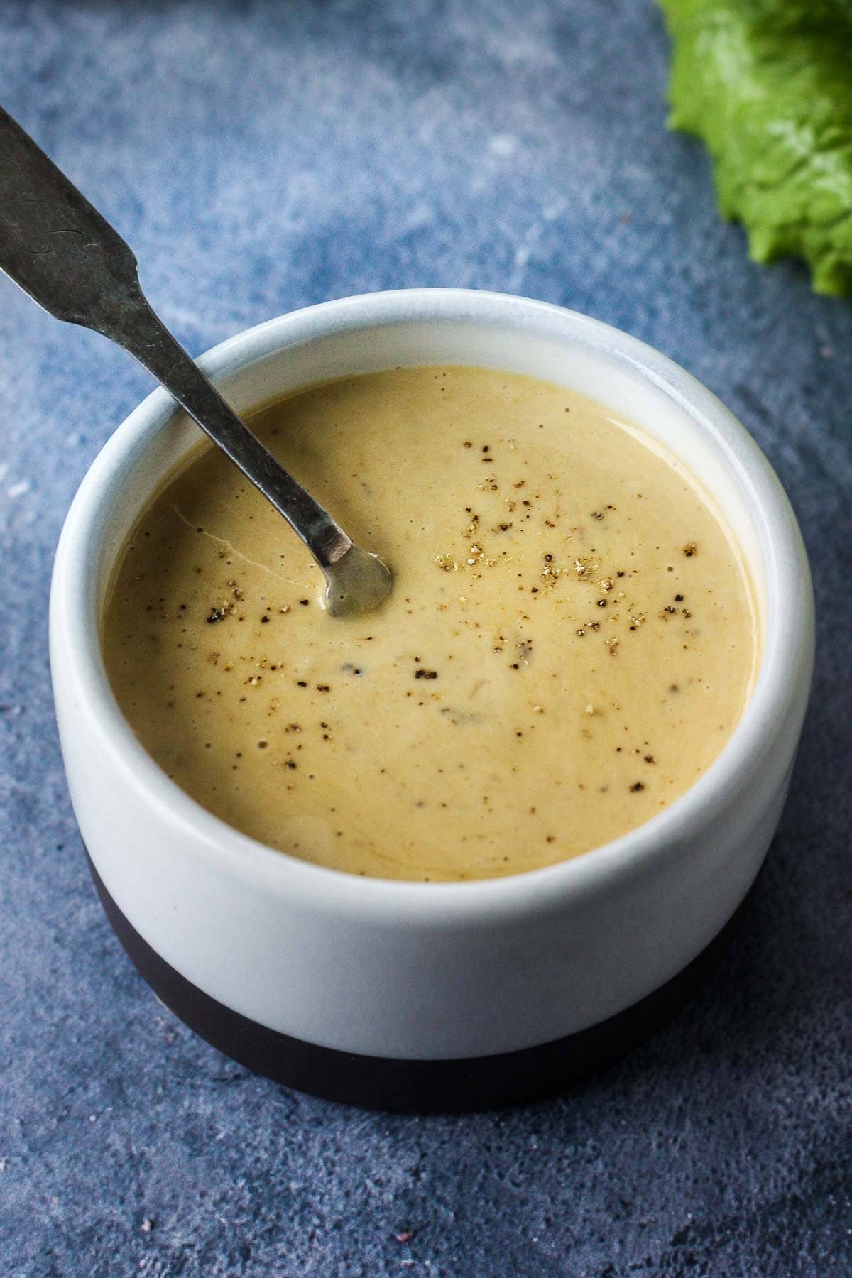 small white bowl with homemade caesar dressing and a silver spoon, topped with black pepper.