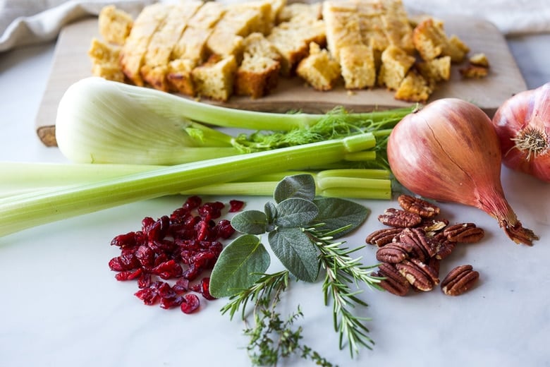 ingredients in stuffing laid out on countertop- wood board with cubed cornbread, fennel, celery, shallots, pecans, fresh herbs, and cranberries.