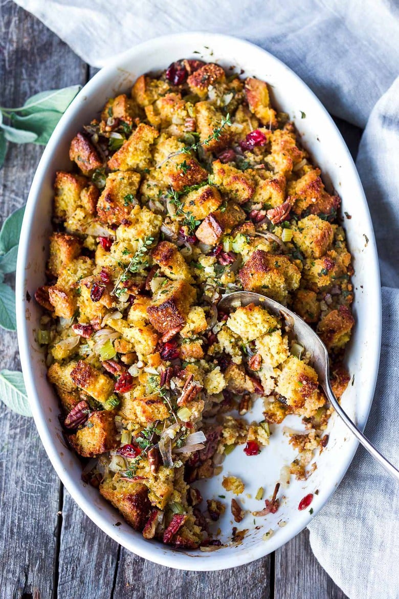 oval baking dish with cornbread stuffing with cranberries, pecans, and herbs, with a serving spoon scooping out some of the stuffing.