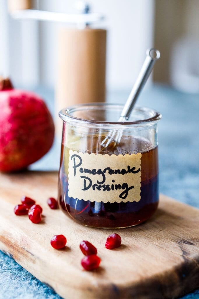 glass jar with pomegranate dressing with label that reads, "pomegranate dressing," with tiny whisk next to pomegranate seeds. 