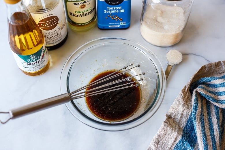 bowl of sesame dressing with whisk in front of dressing ingredients: sesame oil, rice vinegar, mirin, sugar, soy sauce.