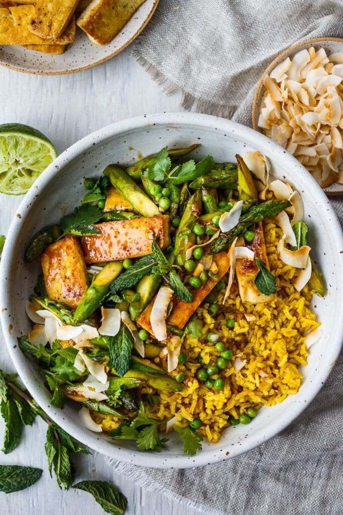 Delicious Coconut Rice Bowl is healthy and adaptable!  Curried jasmine rice cooked in coconut milk and topped with tofu or your choice of protein and seasonal vegetables.  All tossed with a simple, flavorful sauce, fresh cilantro, mint and toasted coconut flakes.