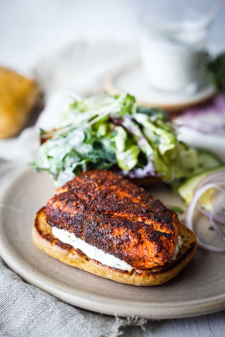 Blackened Salmon Sandwich!  A succulent crispy crusted, cajun spiced, salmon fillet tucked into a toasted bun with fresh greens, cucumber, red onion, drizzled with creamy Dilly Ranch Dressing.