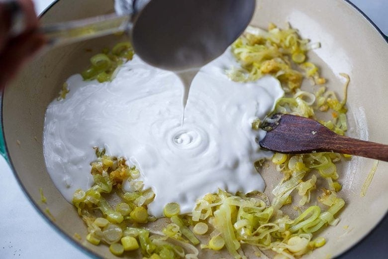 pouring coconut milk from a measuring cup into a braiser of sautéed leeks.