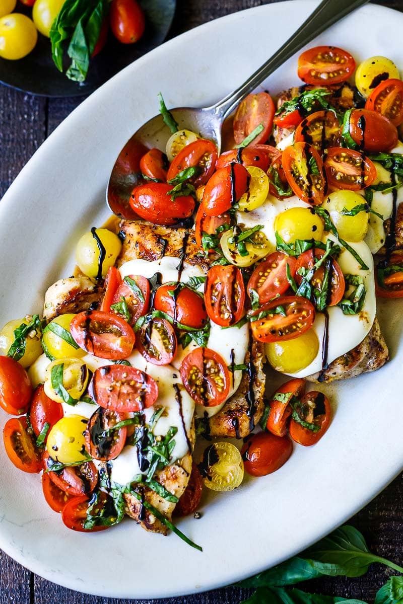 serving plate with grilled chicken caprese topped with mozzarella, cherry tomatoes, sliced basil, and a drizzle of balsamic glaze.