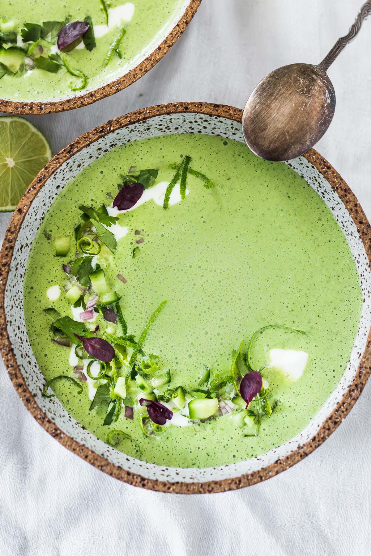 Cucumber Gazpacho in a white speckled bowl with cream and herbs swirled on top.