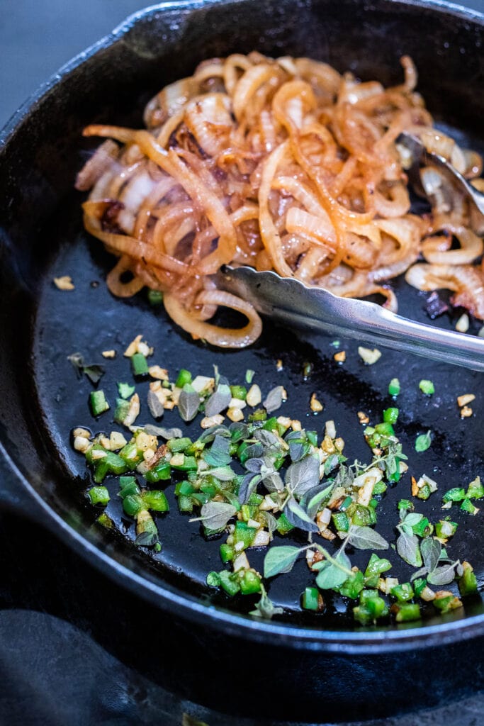 adding jalapeños, oregano, and garlic to cast iron skillet with caramelized onions and tongs.