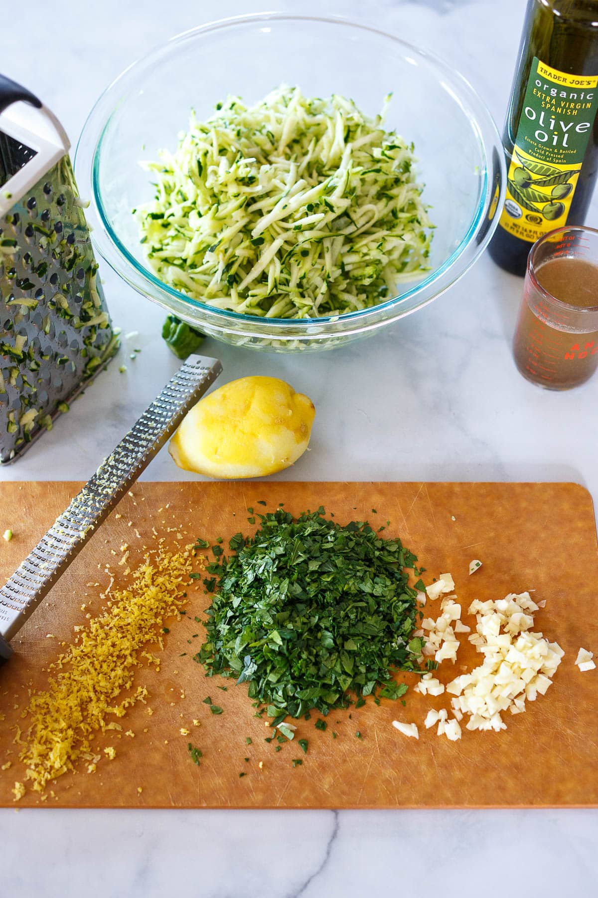 Chopped lemon zest, parsley and garlic on a cutting board with grated zucchini in a glass bowl.