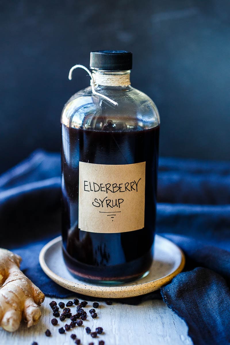 homemade elderberry syrup in glass jar with label, next to ginger root and dried elderberries.