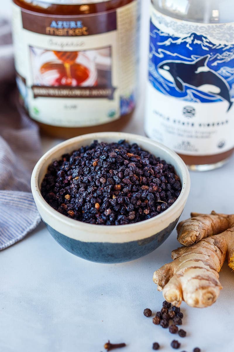 Dried elderberries shown in a blue and white ceramic bowl with apple cider vinegar , honey and fresh ginger in the back ground.