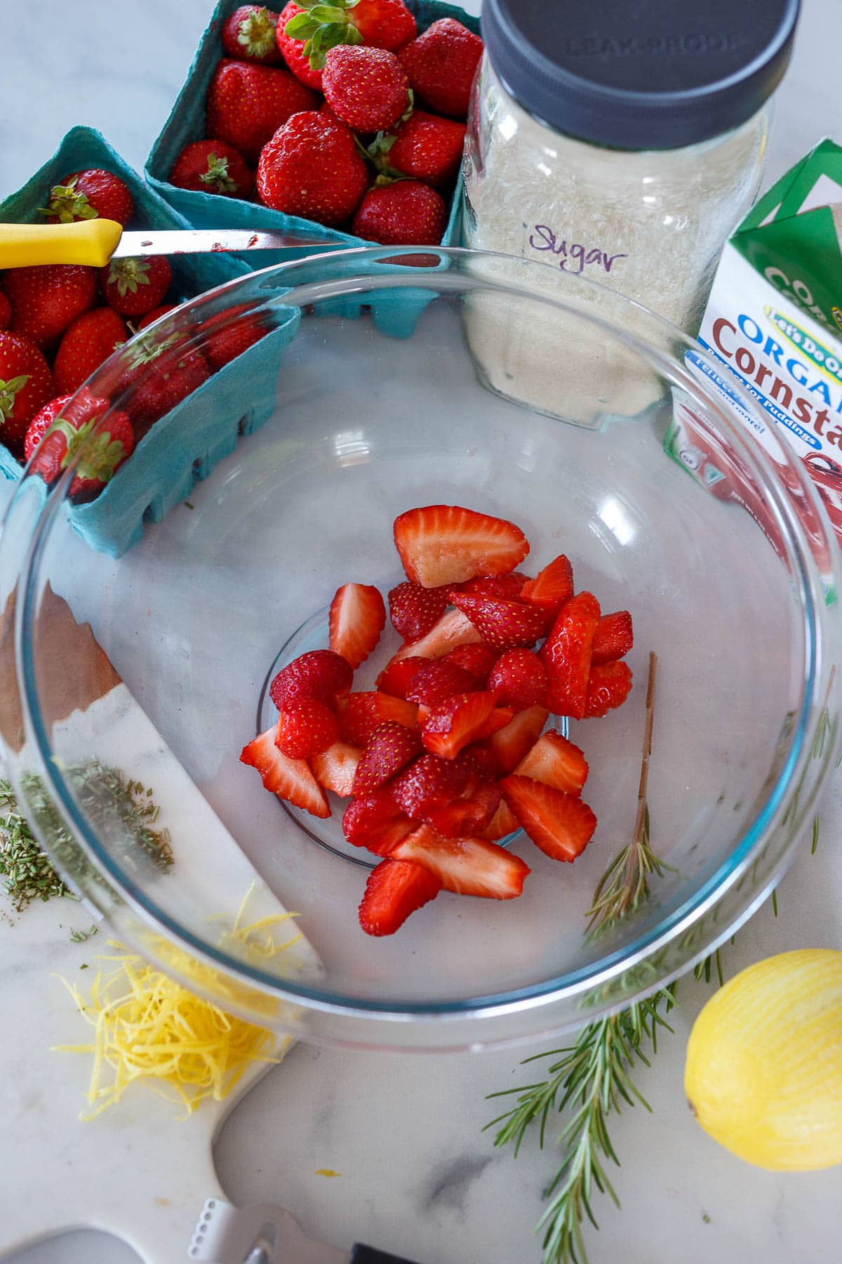 A glass bowl of strawberries with sugar, cornstarch, rosemary and lemon zest around it.