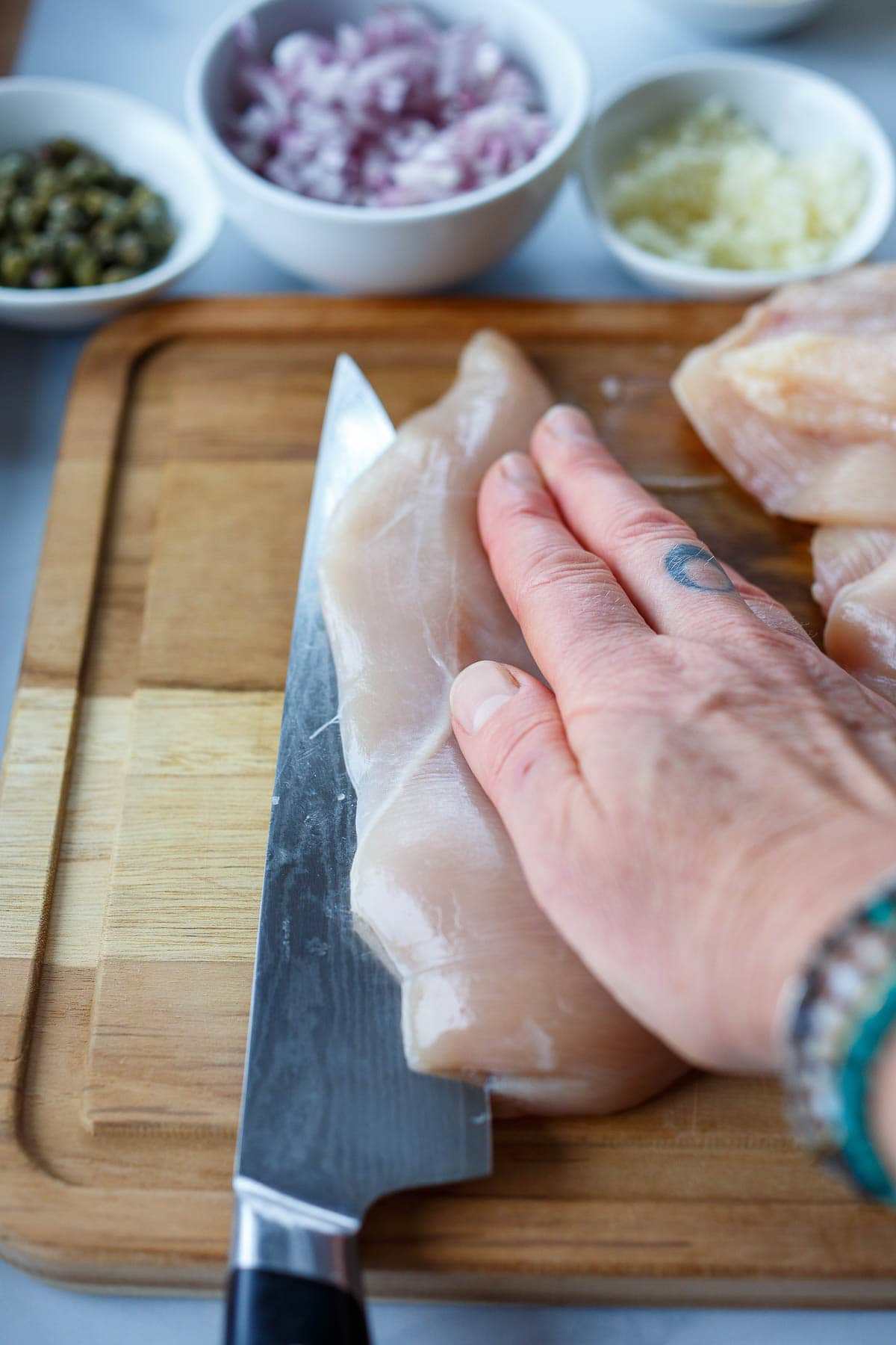 Slicing the chicken breast in half lengthwise on a wooden cutting board.  Bowls of capers, shallots and garlic.