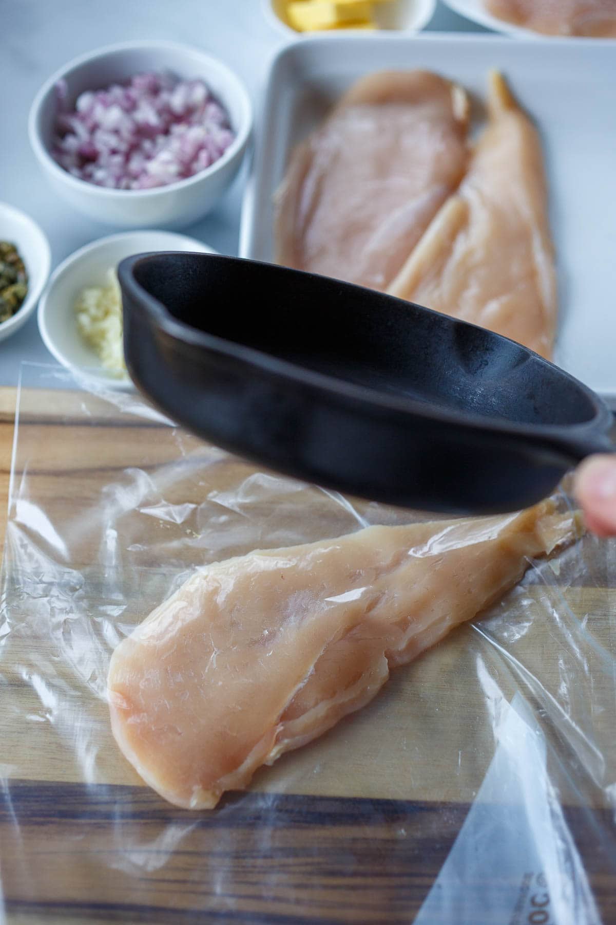 Pounding the chicken on a wooden cutting board, covered in plastic, with a small cast iron skillet.