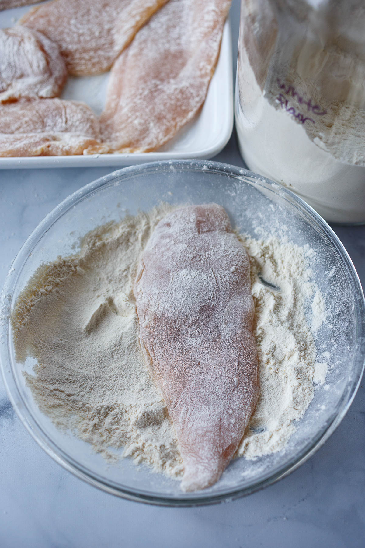 A bowl of flour, lightly dredging chicken cutlet in the flour.