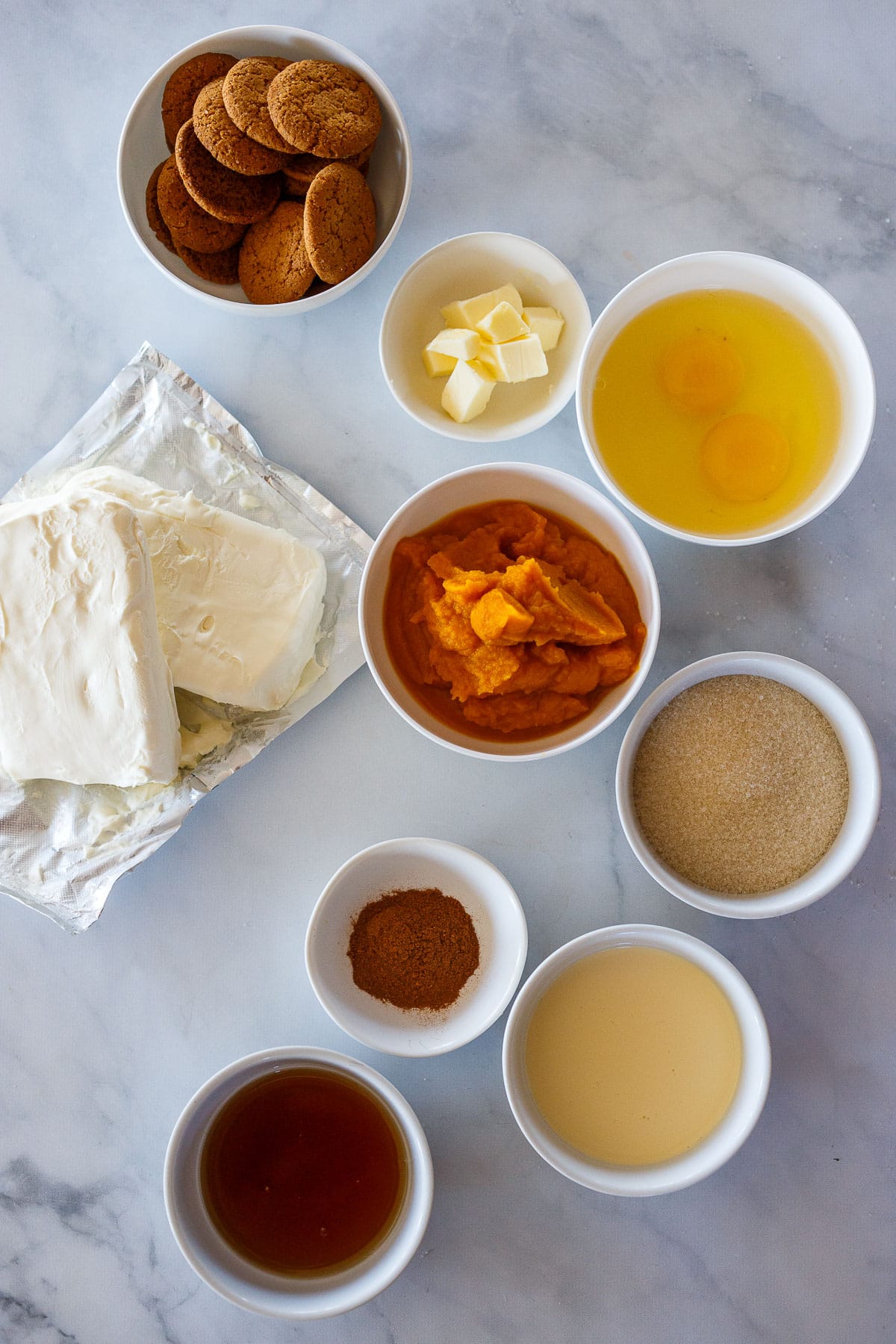 Individual recipe Ingredients for cheesecake in white bowls on marble background.