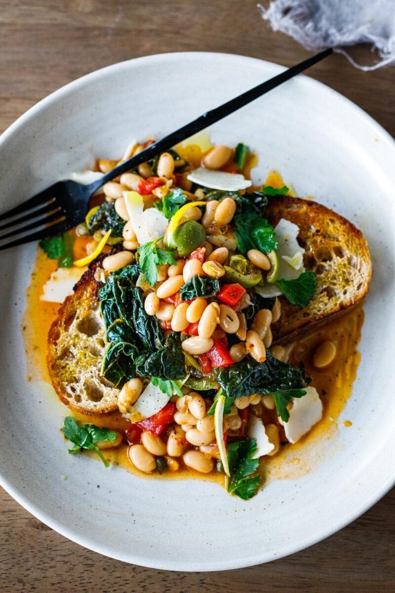 Brothy Beans with lacinato kale, roasted peppers, olives, capers and lemon zest over garlic-infused  Sourdough Bread -a simple pantry meal that can be made in under 30 minutes! Vegan adaptable.