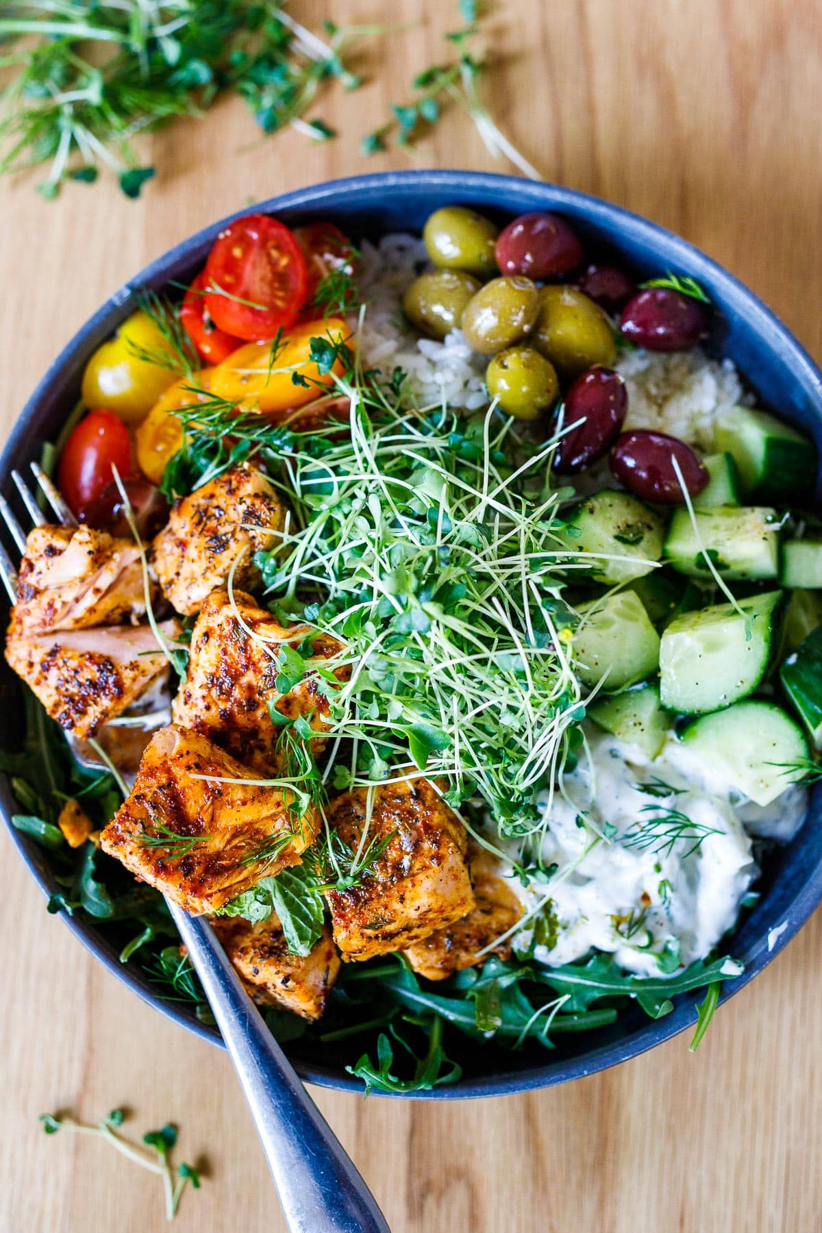 Delicious and healthy Greek Salmon Bowl with rice, greens, olives, tomatoes, cucumber, tzatziki and greek dressing.