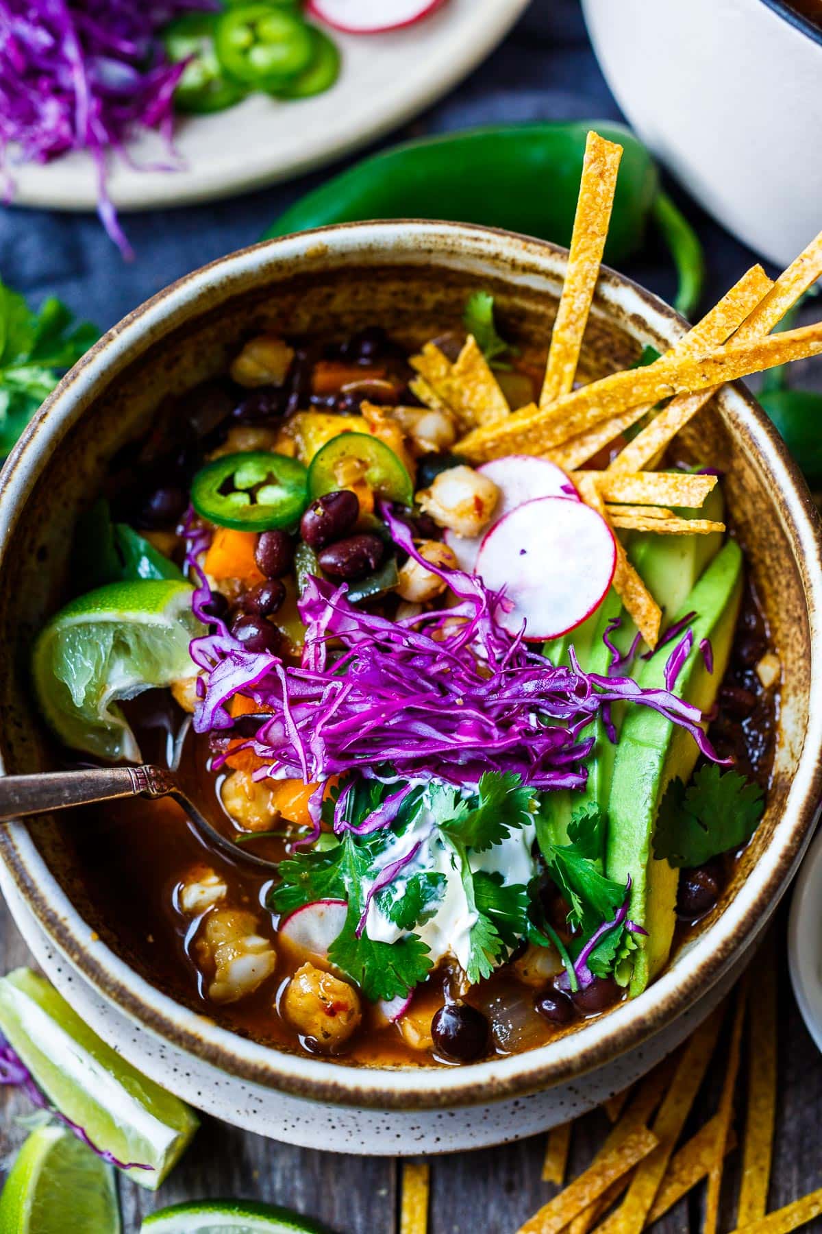 Vegan pozole in a bowl with all the colorful toppings- purple cabbie, cilantro, avocado, strips of tortillas, radishes, lime. 