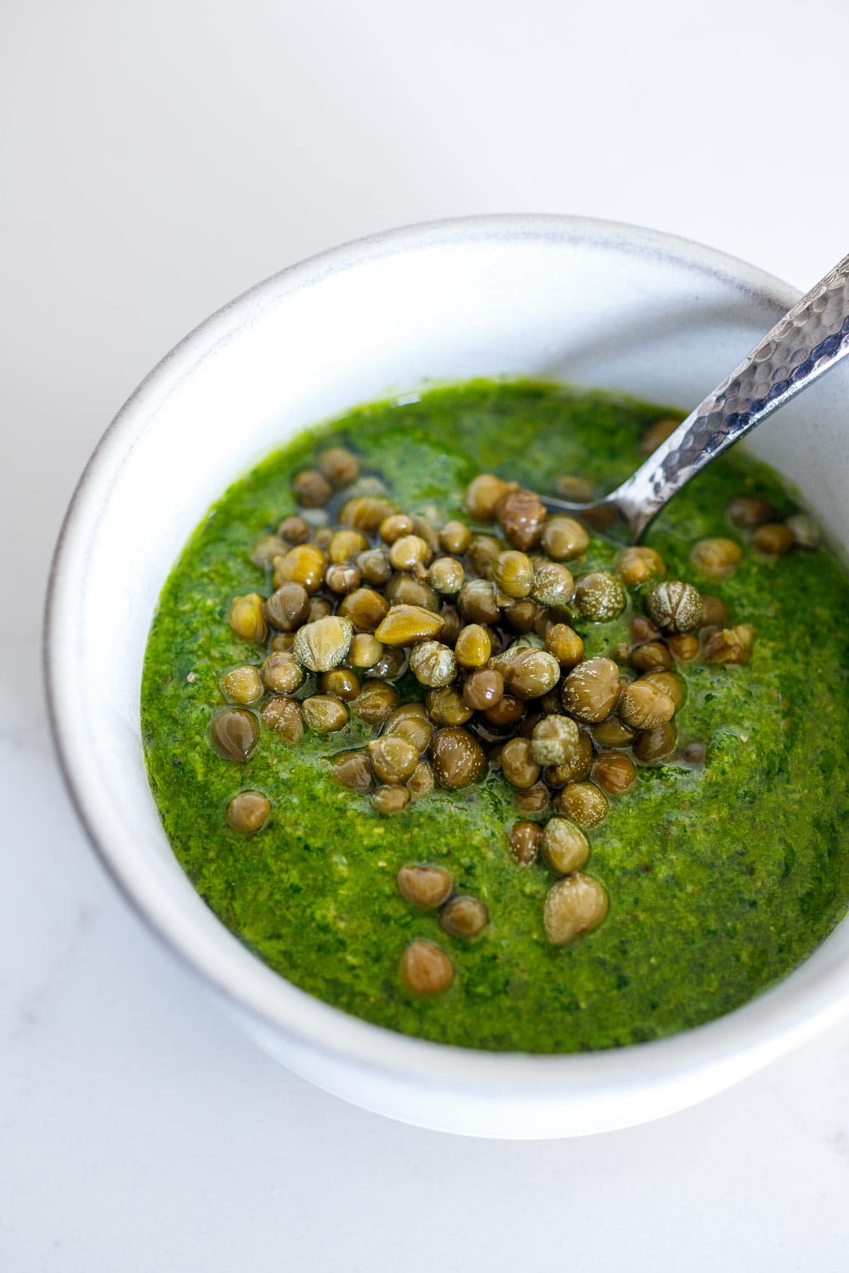 Salsa Verde in a bowl with capers,being stirred in. 