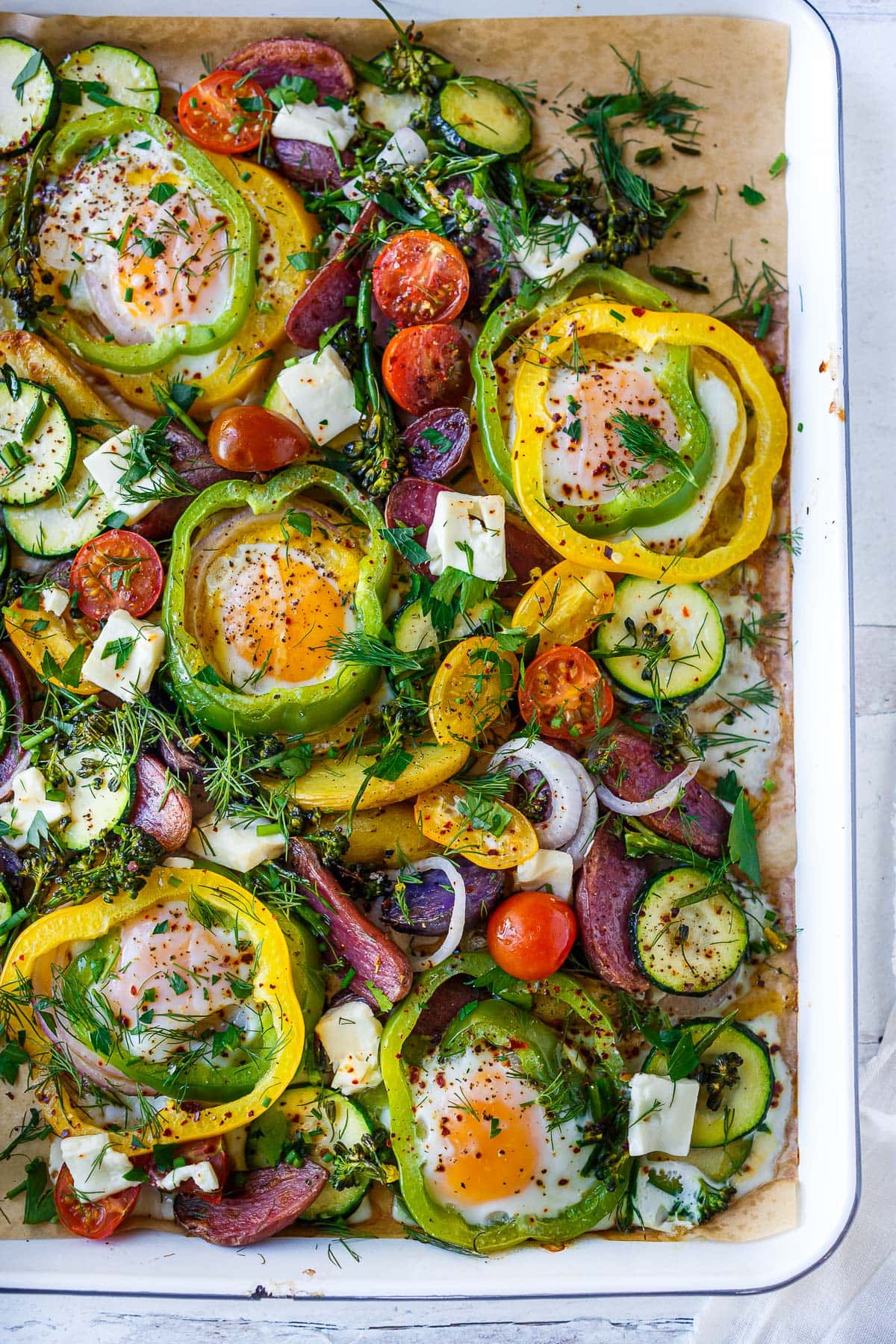 These Baked Eggs are so fun and easy to make! Eggs are baked in the oven on a sheet pan cradled inside rings of bell pepper along with roasted potatoes, zucchini, and tomatoes. Top them with feta and fresh herbs. 