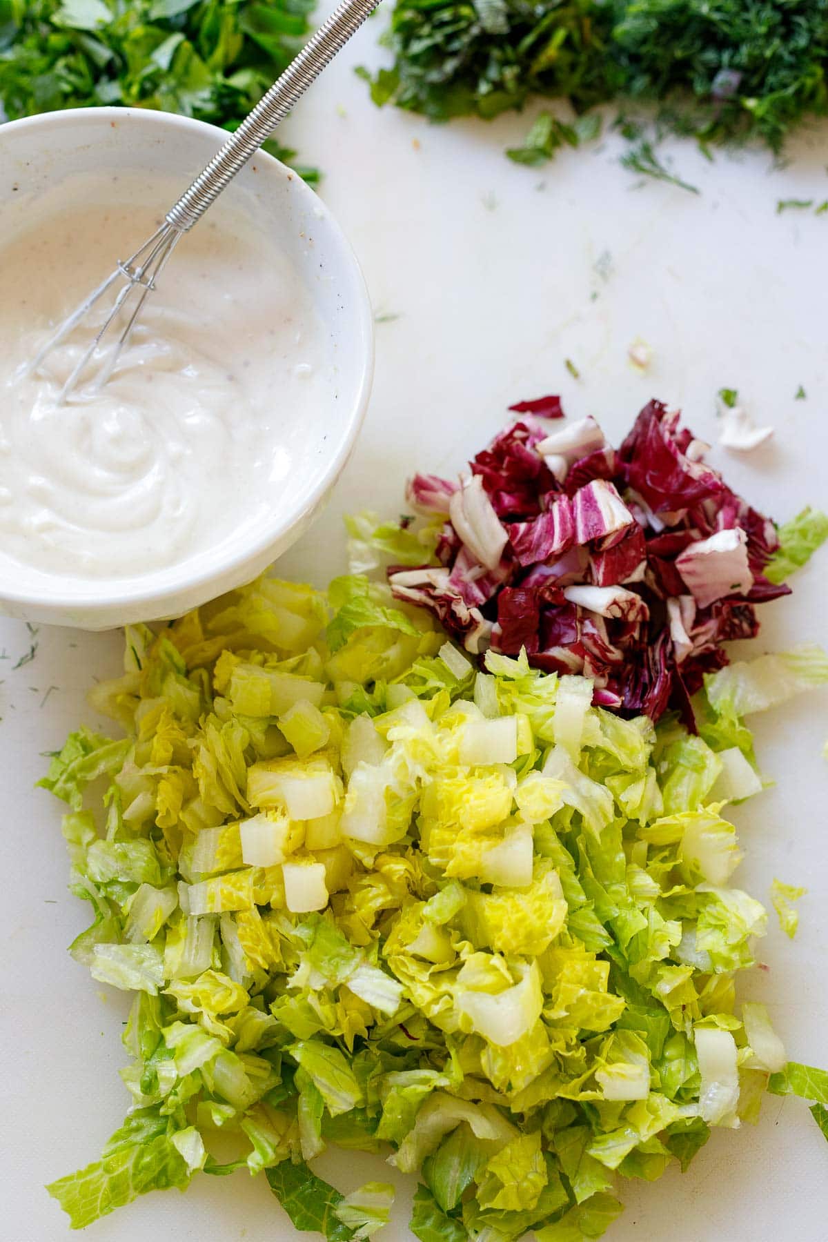 Yogurt tahini sauce in a bowl with a mini whisk and chopped lettuce on a cutting board. 