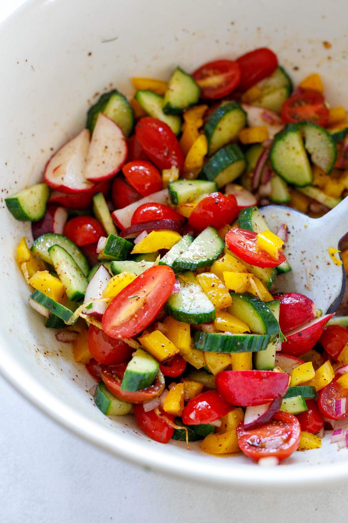 Vegetables and herbs being tossed with dressing in a white bowl.