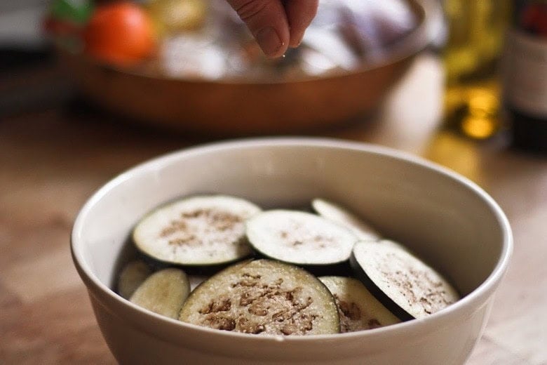 fingers adding a pinch of salt to a bowl of sliced eggplant.