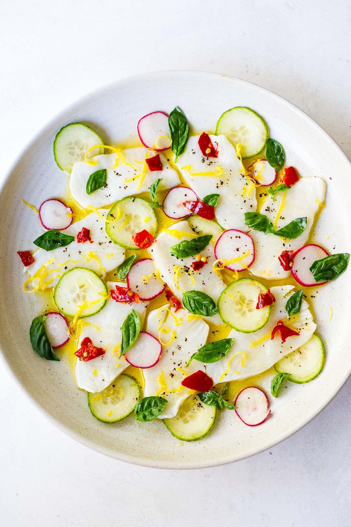 hamachi crudo on white plate with sliced cucumber, radish, chili peppers, lemon zest and basil