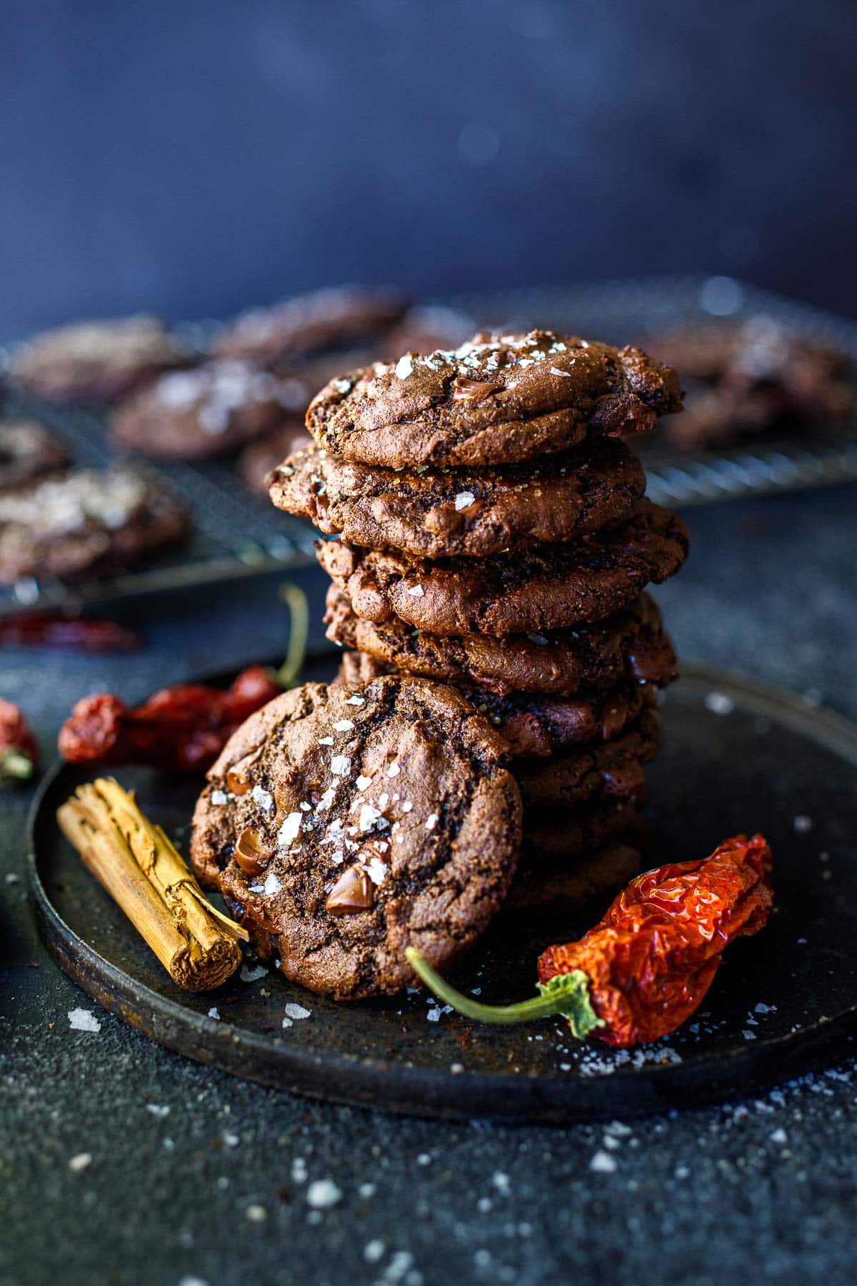 stack of double chocolate chip cookies on plate with flaky salt and dried cayenne pepper and cinnamon stick on side