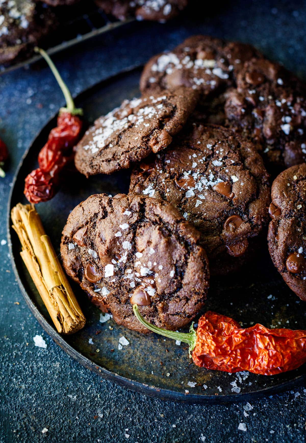 Plate of double chocolate chip cookies with flaky salt and dried cayenne peppers and cinnamon stick on side