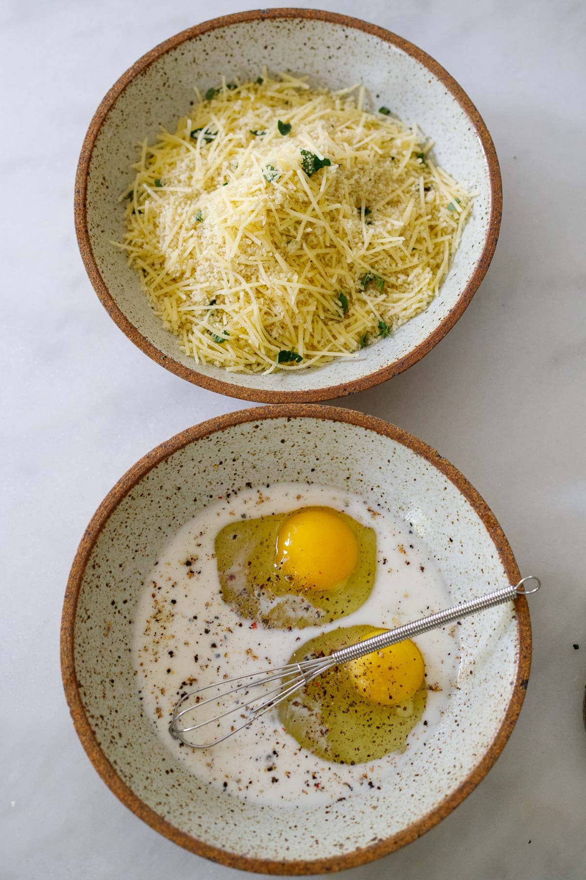 two ceramic bowls, one with egg wash and whisk, one with parmesan and breadcrumbs