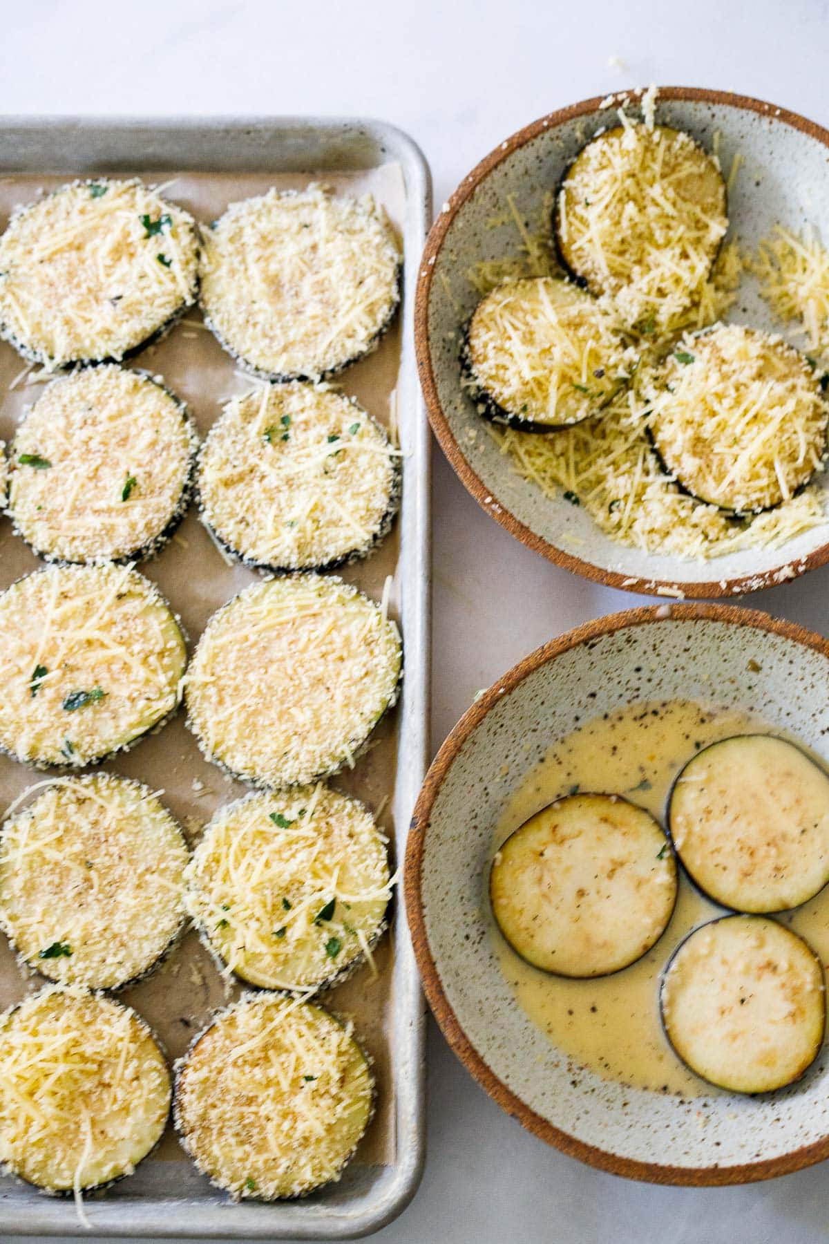 eggplant slices in egg wash bowl, in parmesan breadcrumb bowl, and arranged on baking sheet
